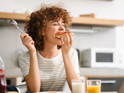 Deine Ernährung. Dein Kurs. Unser Zuschuss. Eine junge Frau sitzt am Küchentisch und beißt in ein Brot mit Marmelade.