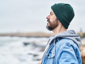 Mann mit Mütze steht mit geschlossenen Augen am Meer 