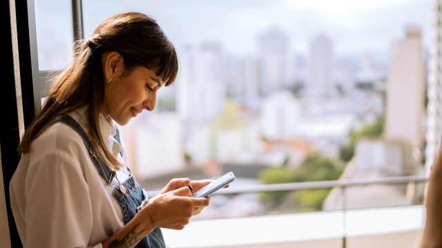 Eine junge Frau steht am Fenster und schaut auf ihr Handy