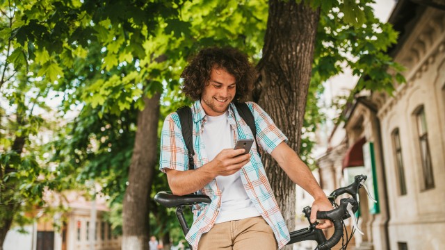 Ein junger Mann steht an seinem Fahrrad und schaut auf sein Handy. 