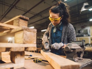 Eine junge Frau schleift ein Stück Holz in einer Werkstatt. 