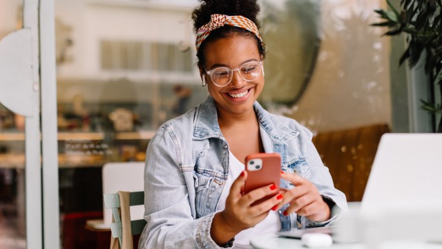 Eine Frau sitzt im Cafe mit Laptop und schaut auf ihr Smartphone